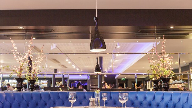 Un comedor con asientos tapizados en azul, una mesa redonda con copas de vino y cubiertos, y jarrones decorativos con flores.