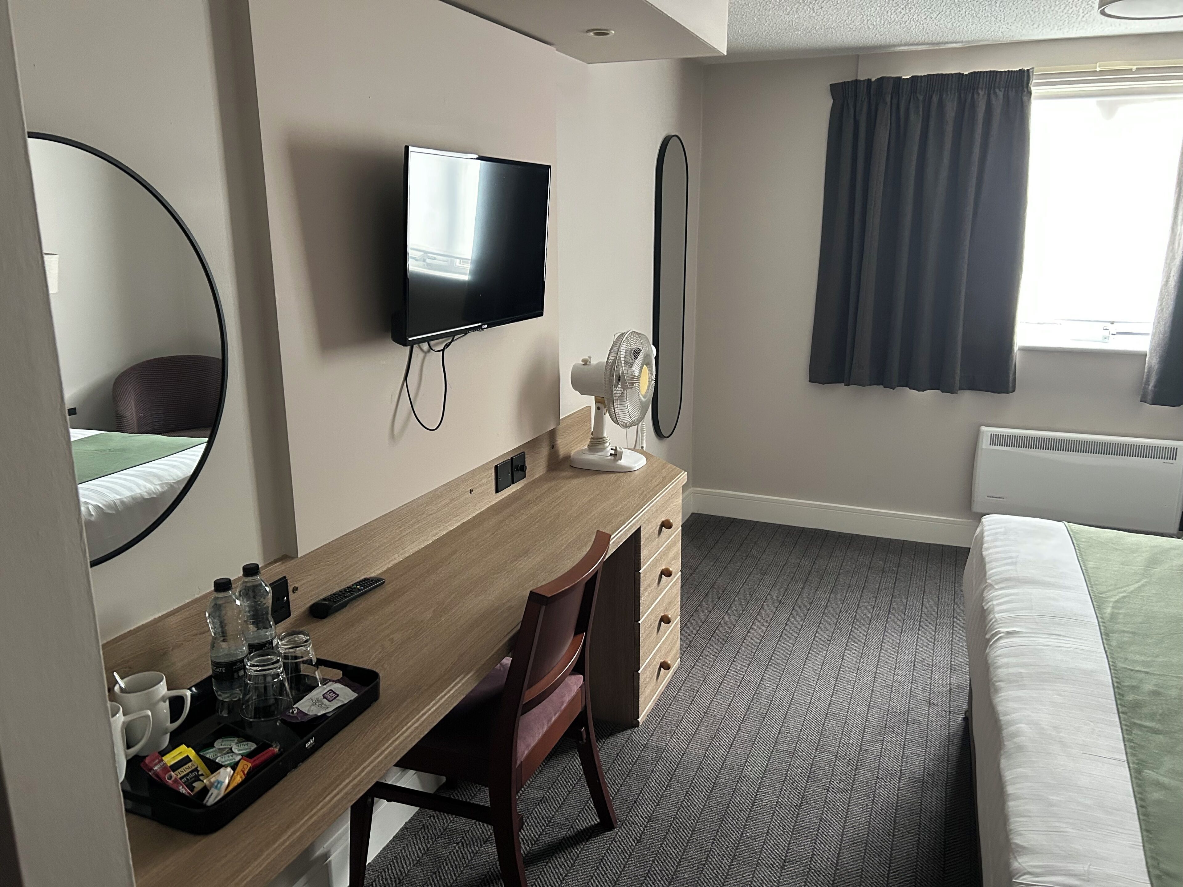 Quarto de hotel com cama, escrivaninha, cadeira, televisão, espelho, ventilador e janela com cortinas.