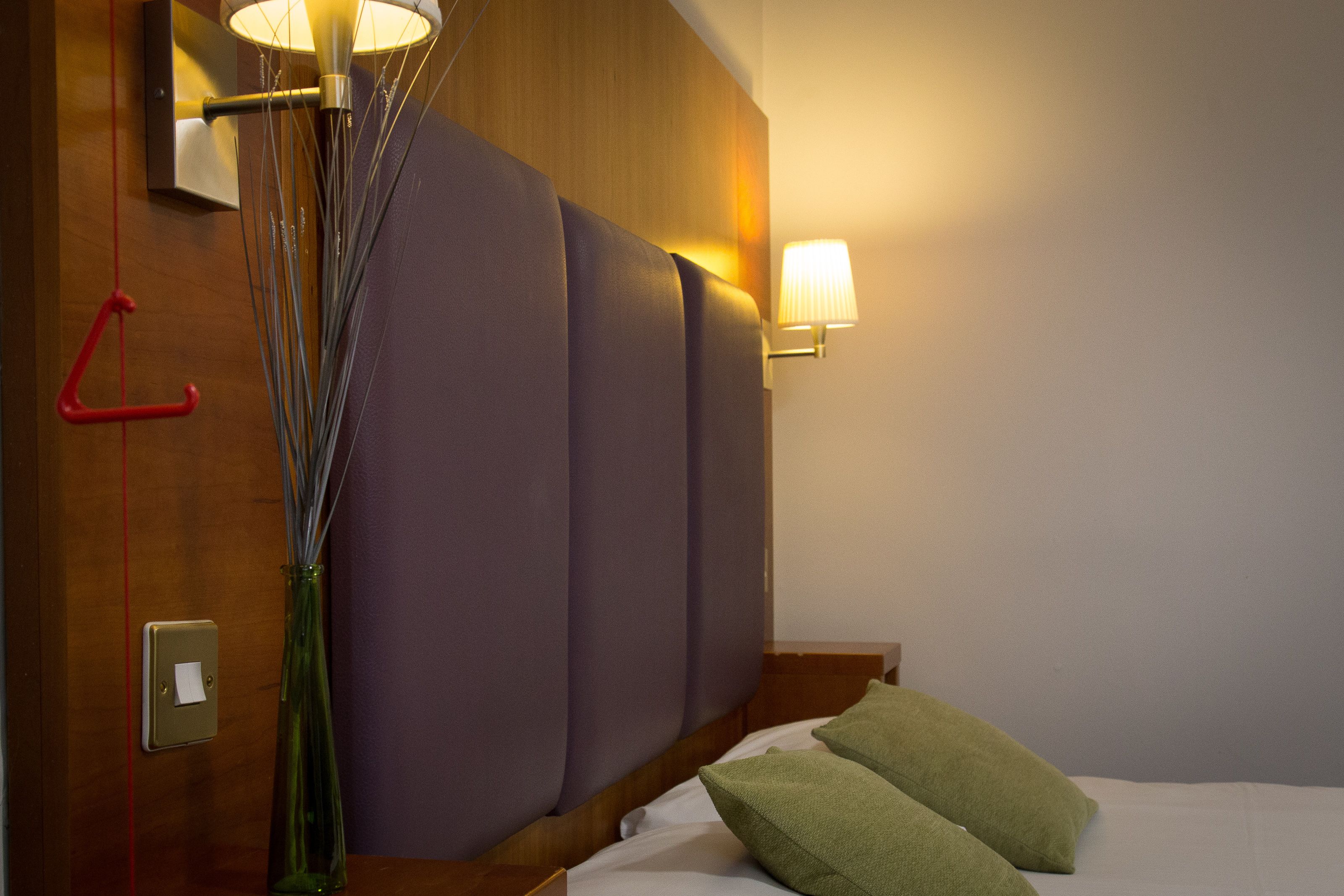 A hotel room with a bed, a nightstand, a lamp, and a vase with a red hanger.