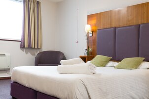 Una habitación de hotel con una cama grande, una silla, una mesita de noche con lámpara y un cabecero de madera.