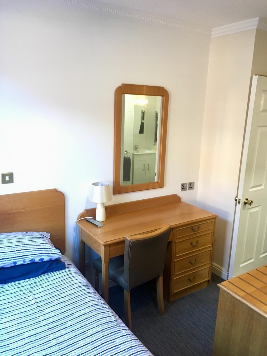 Habitación de hotel con cama, escritorio con silla, cómoda, espejo y puerta.