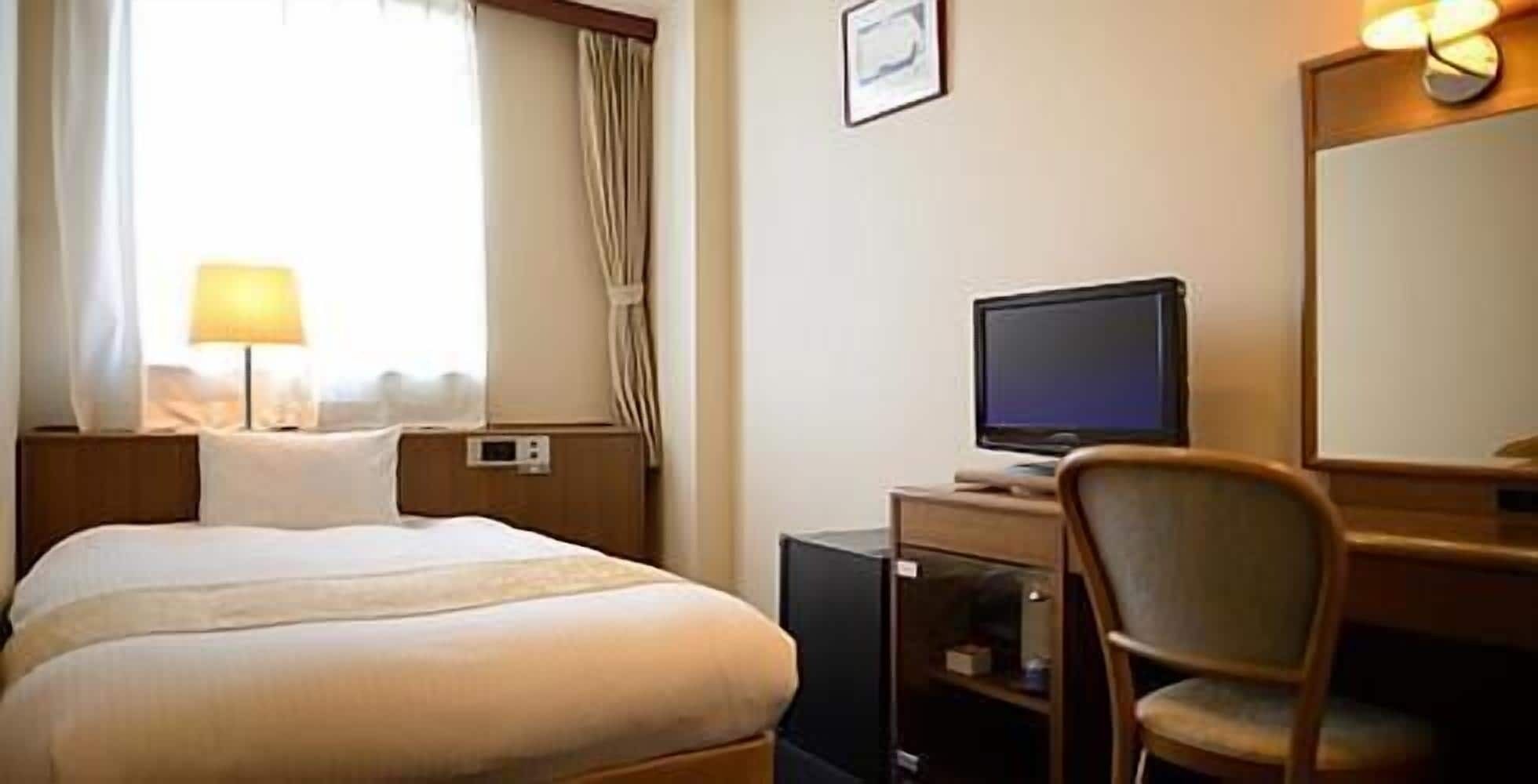 A hotel room with a bed, a desk with a computer, a chair, a lamp, and a window with curtains.