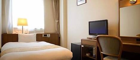 A hotel room with a bed, a desk with a computer, a chair, a lamp, and a window with curtains.