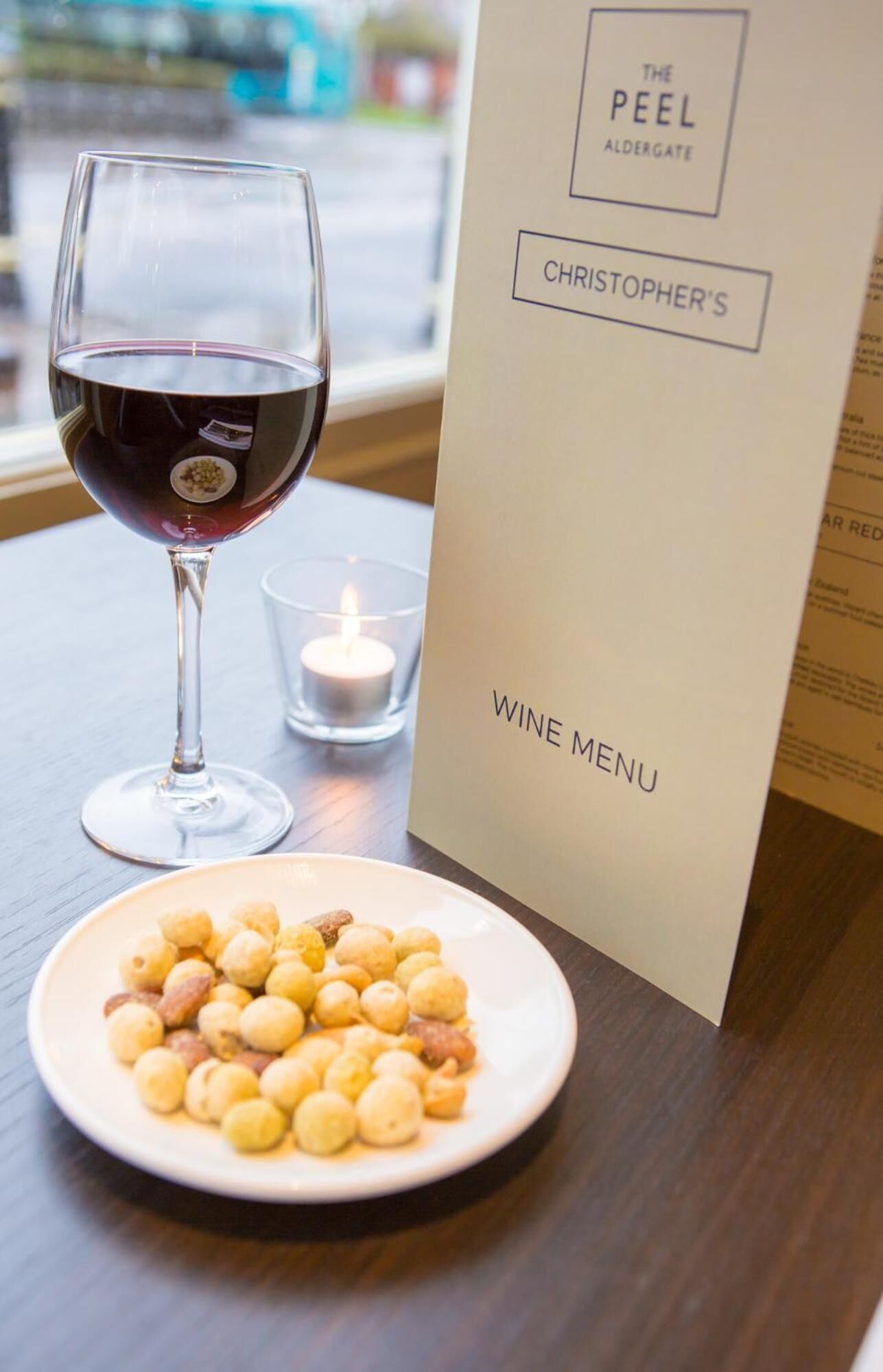 A wine glass with red wine, a candle, and a plate of nuts on a table with a menu titled "The Peel Aldergate Christopher's Wine Menu".
