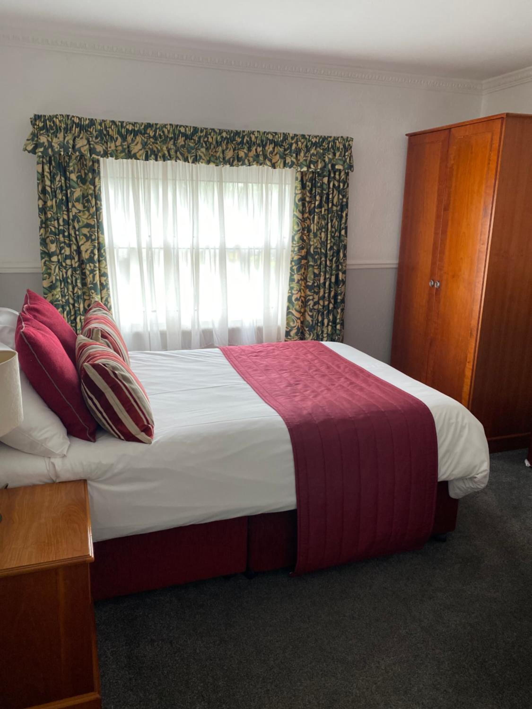 A neatly made bed with a white and red bedspread, a wooden wardrobe, and a window with floral curtains.