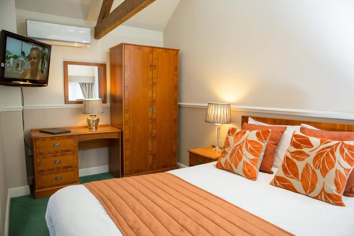Quarto de hotel com cama, escrivaninha de madeira, guarda-roupa, televisão e mesa de cabeceira com abajur.