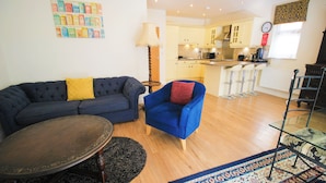 Un salon avec un canapé bleu foncé, un fauteuil bleu et une table ronde. Au fond, on aperçoit une cuisine avec des meubles blancs et une horloge au mur.
