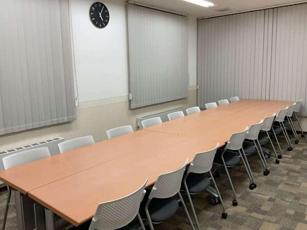Una sala de conferencias con una larga mesa y sillas, un reloj en la pared y persianas en las ventanas.