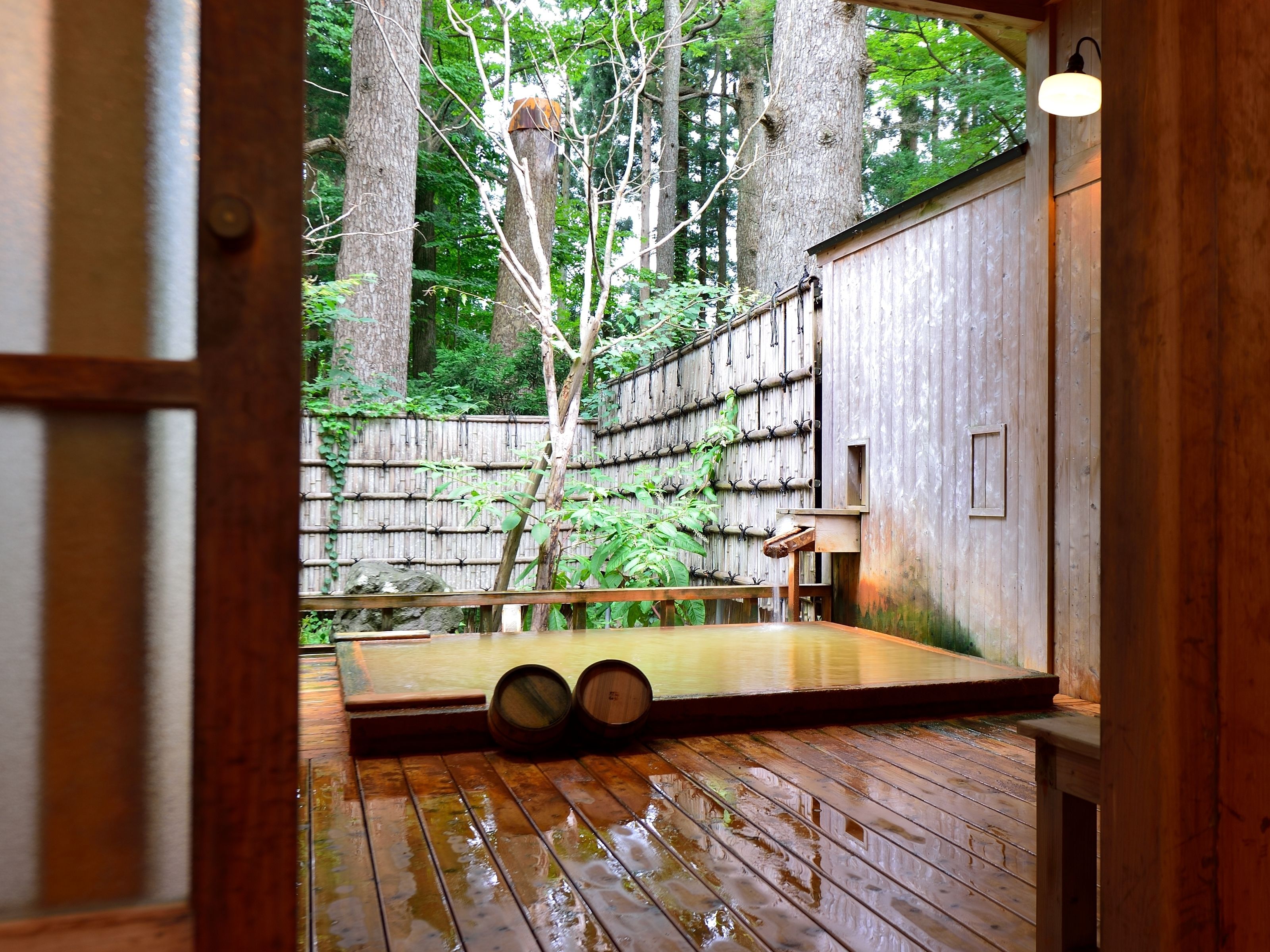 Un deck de madera con un baño de aguas termales, rodeado por una cerca de bambú y árboles.