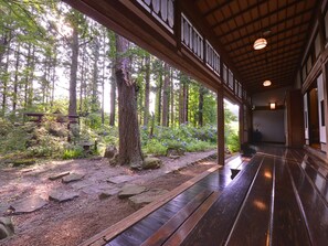 A covered wooden walkway with a stone path, surrounded by a forested area with tall trees and a bench. - Syoho En (Daisen)