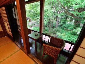 A traditional Japanese room with tatami flooring, a low wooden table, a wicker chair, and a large window offering a view of a lush garden.