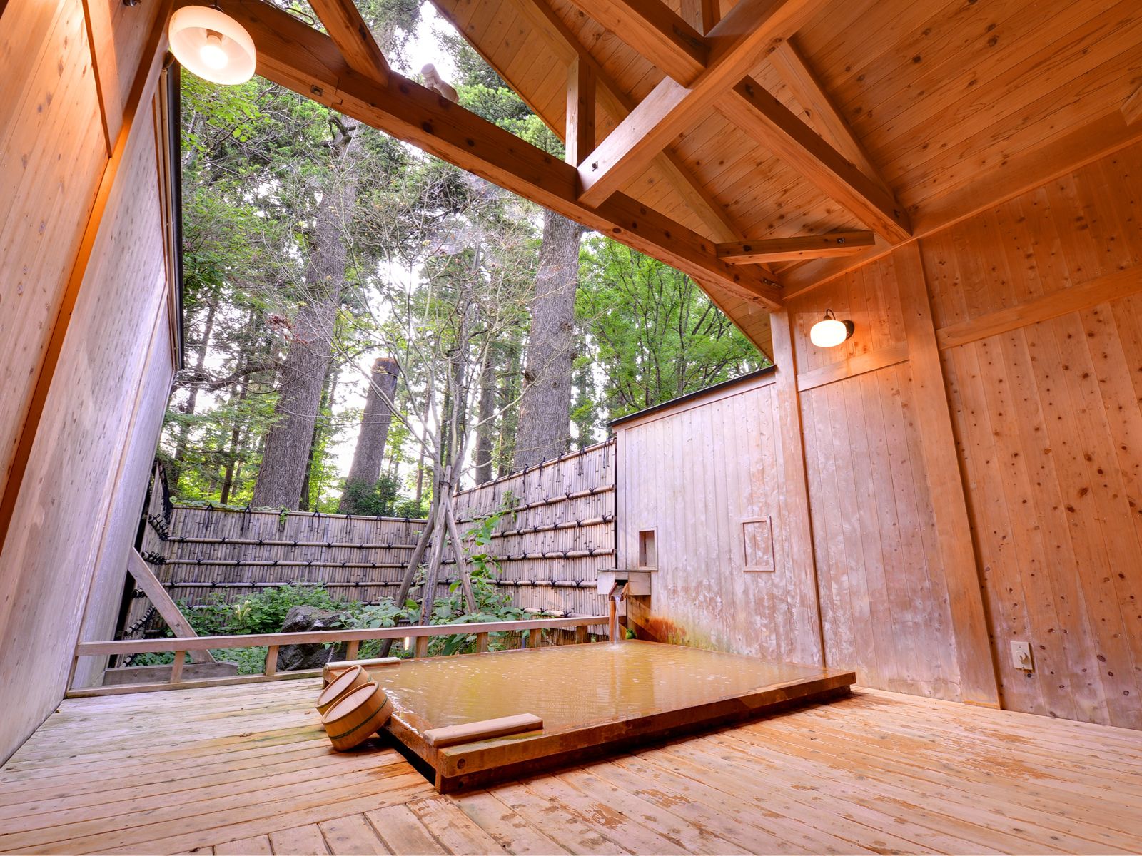 Zona de baños termales de madera con terraza, pared de madera y vistas a altos árboles.