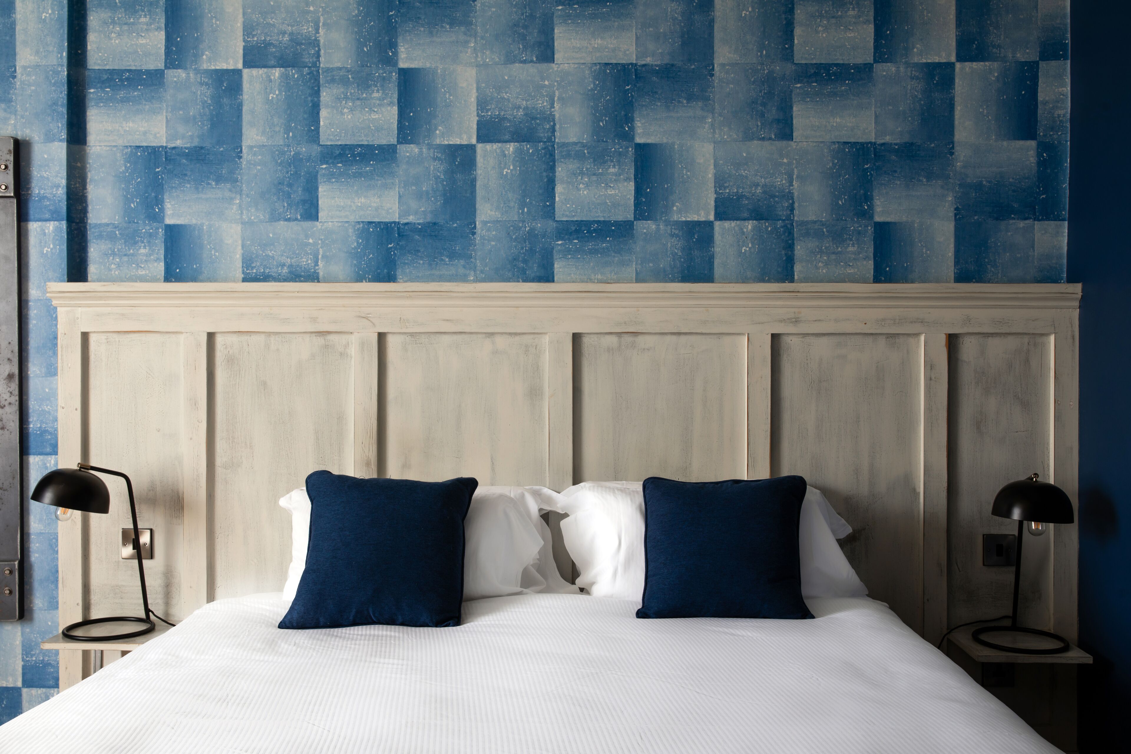 A neatly made bed with white linens and two blue pillows, flanked by two bedside tables with black lamps, against a wall with a geometric blue tile pattern.