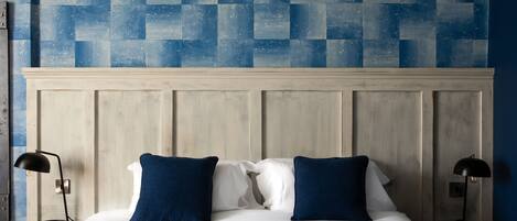 A neatly made bed with white linens and two blue pillows, flanked by two bedside tables with black lamps, against a wall with a geometric blue tile pattern.