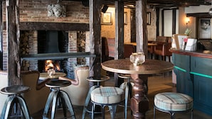 A rustic pub interior with exposed brick walls, a brick fireplace, wooden beams, and a round wooden table with stools. - White Lion Tenterden (Tenterden)