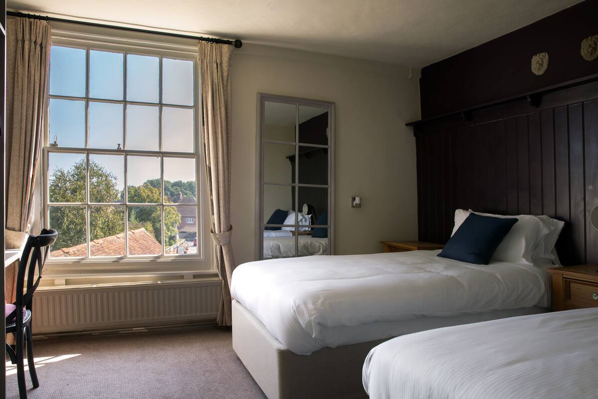 A hotel room with two beds, a large window with curtains, a radiator, a chair, and a wooden headboard.