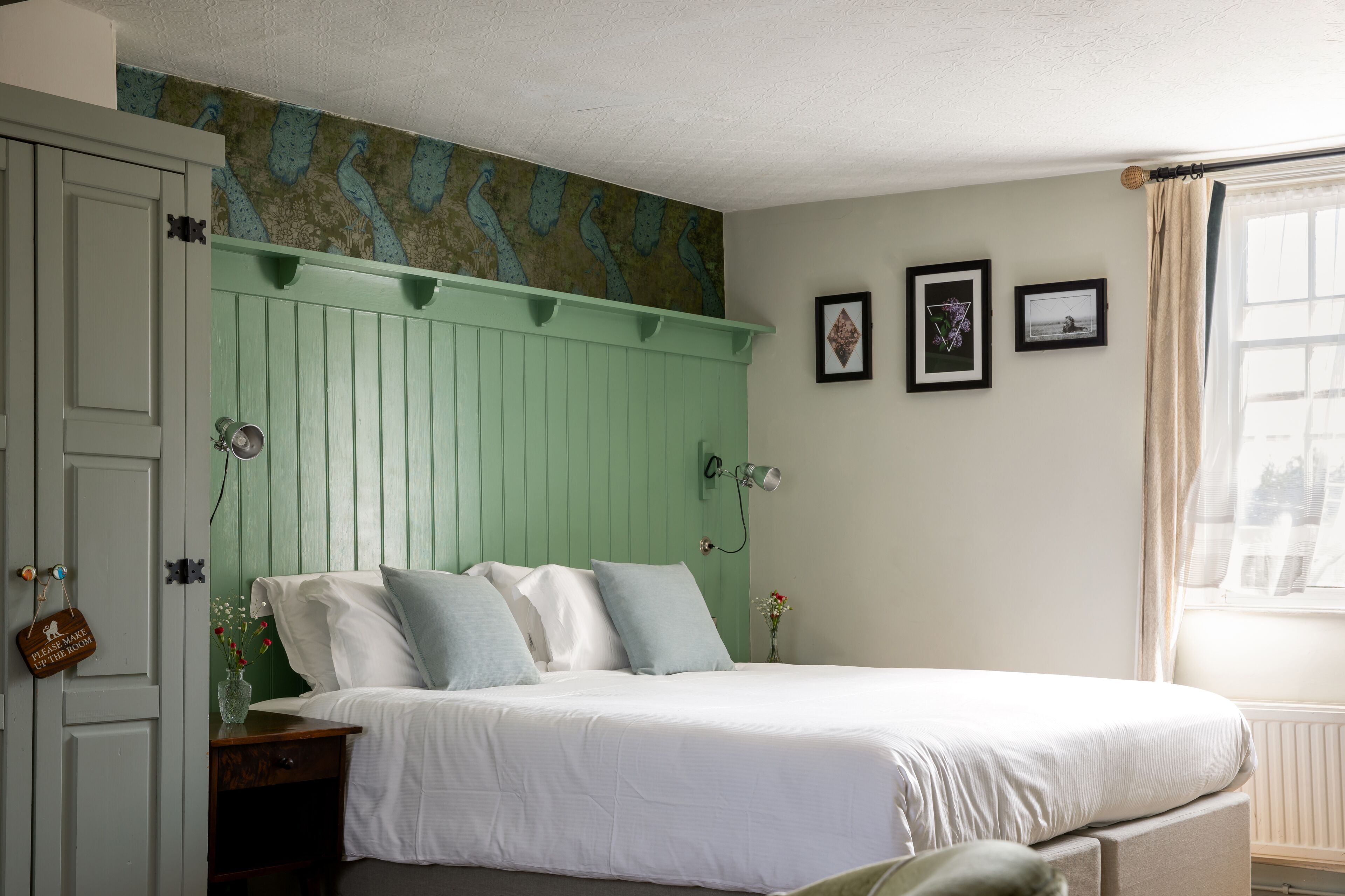 A bedroom with a bed, a nightstand, a wall-mounted lamp, a door with a sign, and a window with curtains.