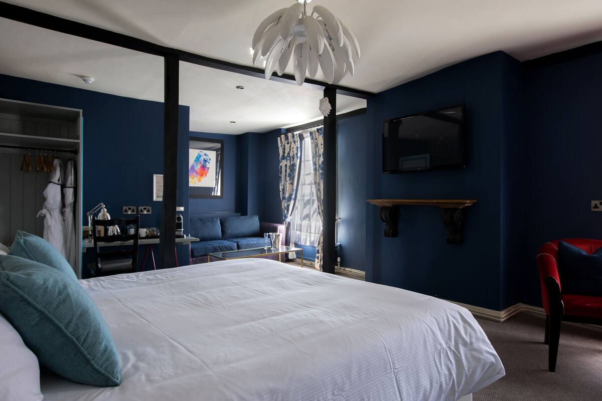 A bedroom with a large bed, a desk with a chair, a red chair, a TV, and a window with curtains.