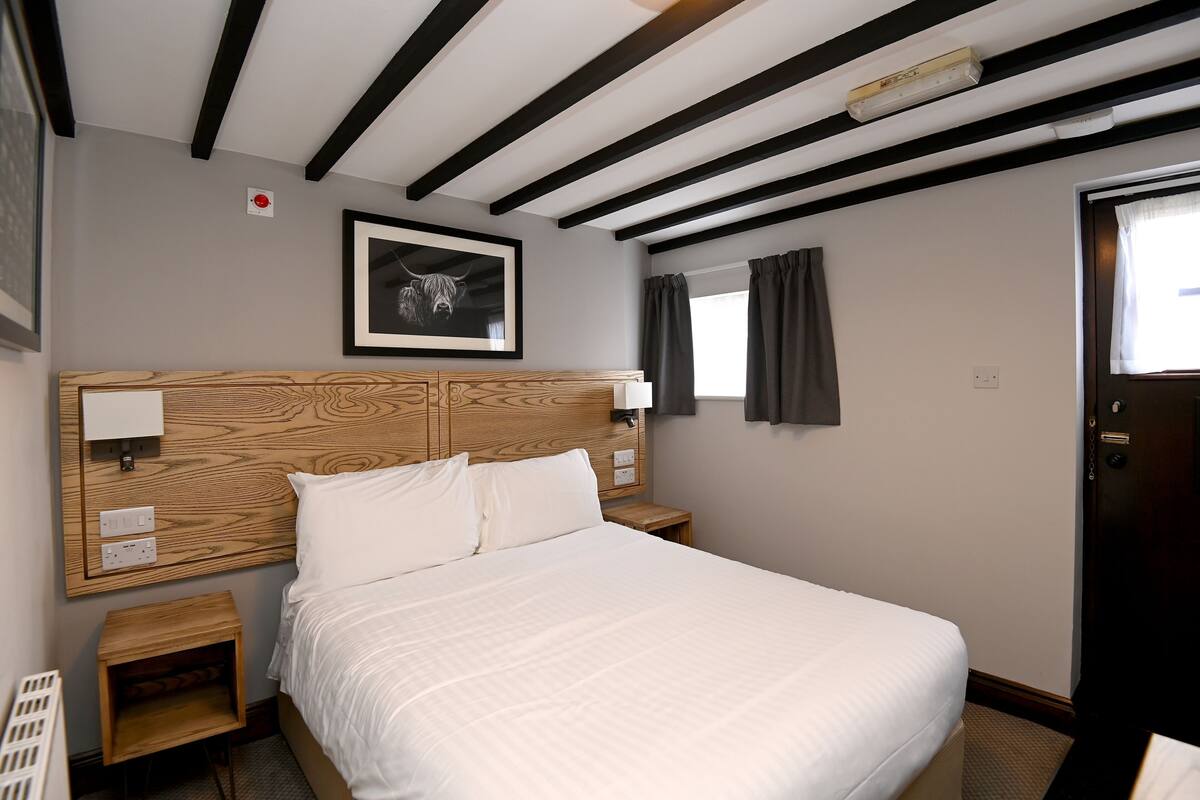 A hotel room with a wooden headboard, a bed with white linens, a framed picture on the wall, a window with curtains, and a door.