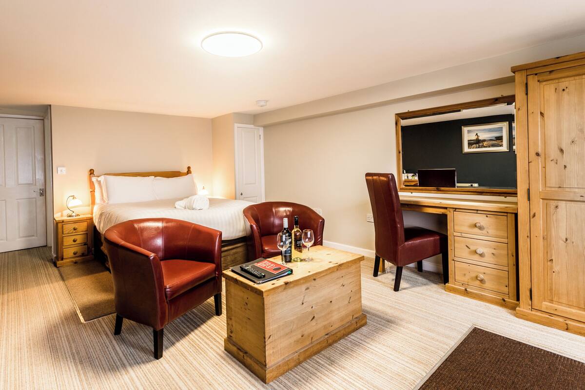 Habitación de hotel con una cama, dos sillones de cuero, un escritorio de madera, un aparador y una mesita con una revista y vasos.