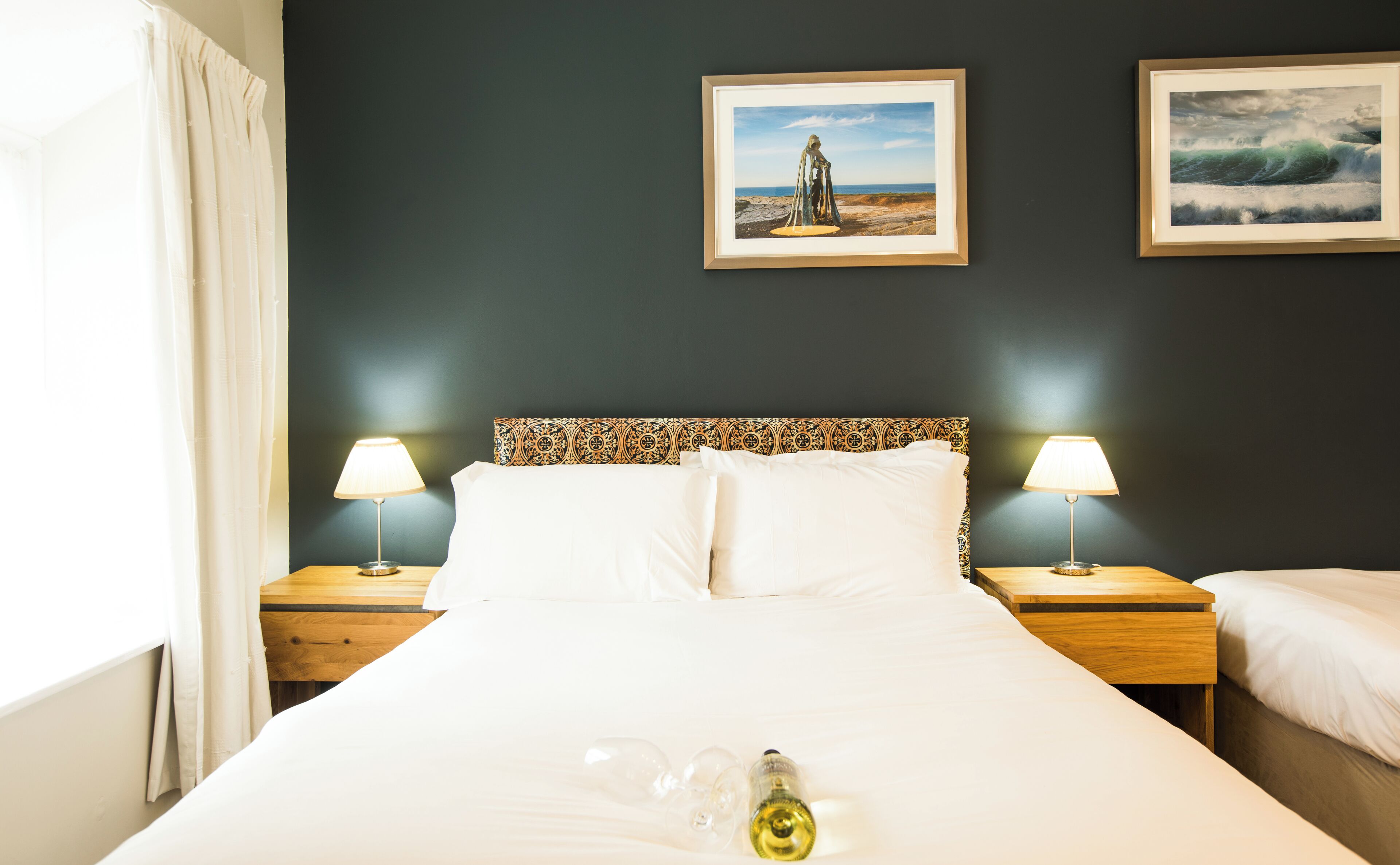 Habitación de hotel con una cama, dos mesitas de noche con lámparas, una ventana con cortinas y dos cuadros en la pared.