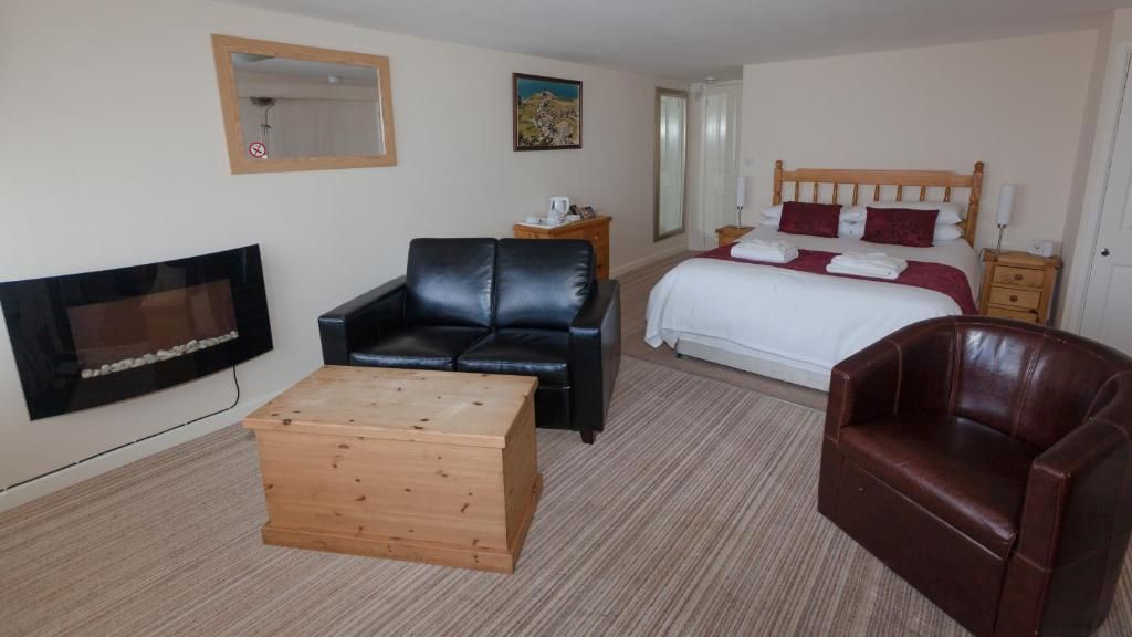 A hotel room with a bed, a black leather sofa, a wooden ottoman, a brown leather armchair, a TV, and a small table.