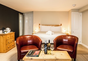 A hotel room with a bed, two leather chairs, a wooden table with wine bottles and glasses, a wooden dresser, and a nightstand with a lamp. - King Arthur's Arms Inn (Tintagel)
