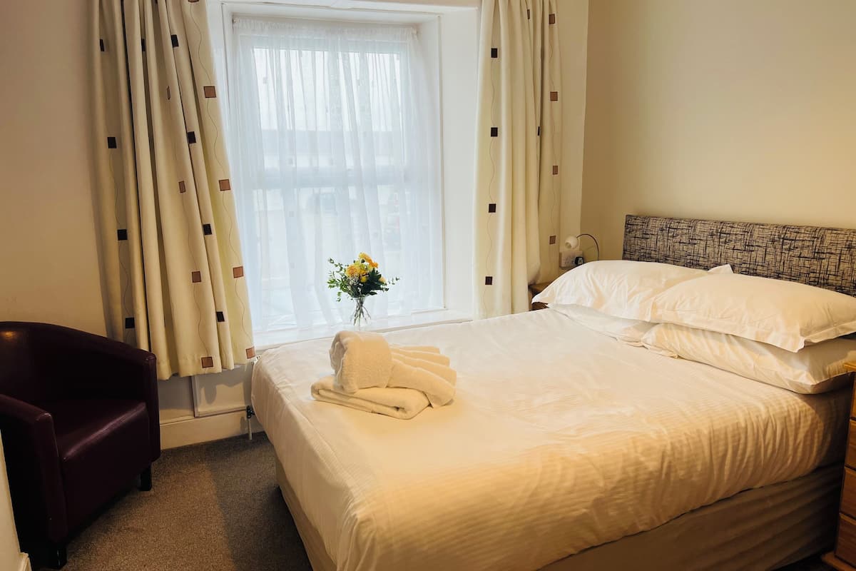 Una habitación de hotel con una cama grande, un sillón oscuro, una ventana con cortinas translúcidas y un jarrón con flores.