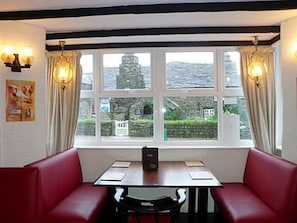 A restaurant with a table and red booth seating, large windows with curtains, and a view of a building outside. - King Arthur's Arms Inn (Tintagel)