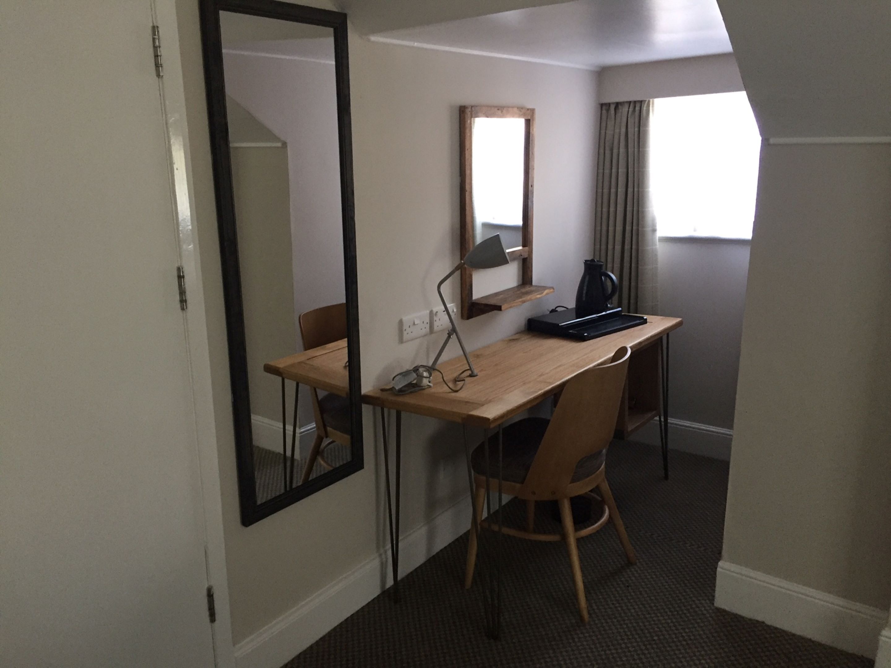 Een kleine hotelkamer met een houten bureau, een stoel, een spiegel, een lamp en een koffiezetapparaat.
