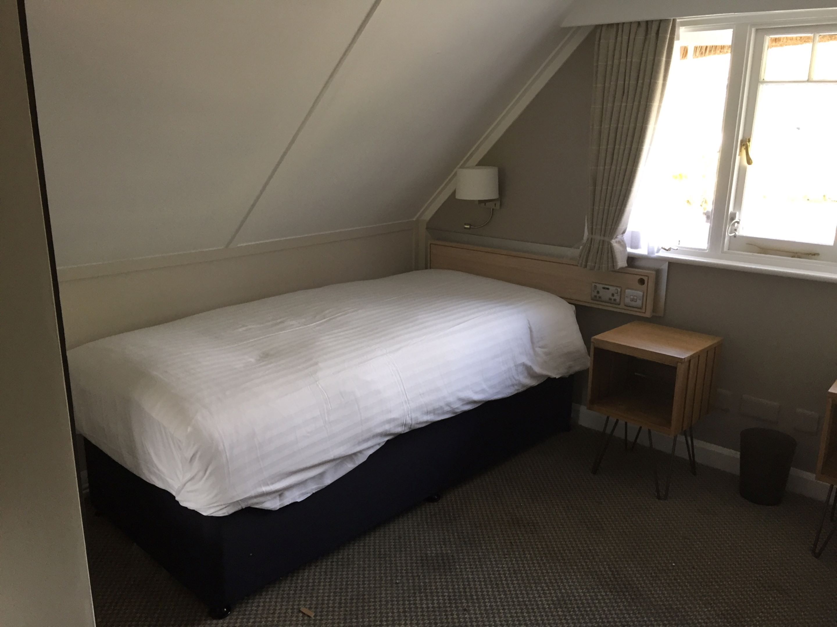 A small, single-bed room with a sloped ceiling, a bedside table, a lamp, and a window with curtains.