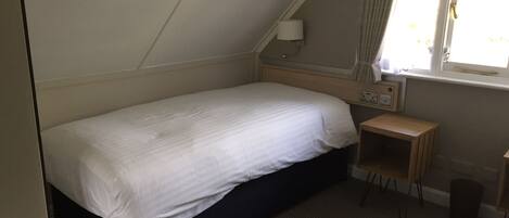 Una habitación pequeña con cama individual, techo inclinado, mesita de noche, lámpara y ventana con cortinas.