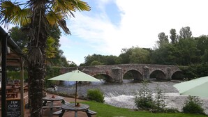 Ein Außenbereich mit Holztischen und -bänken, einer Steinbrücke und einem Fluss im Hintergrund.