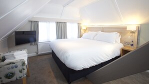 Ein Hotelzimmer mit einem großen Bett, einem wandmontierten Flachbildfernseher, einem Nachttisch mit Lampe und einem mit Vorhängen versehenen Fenster.