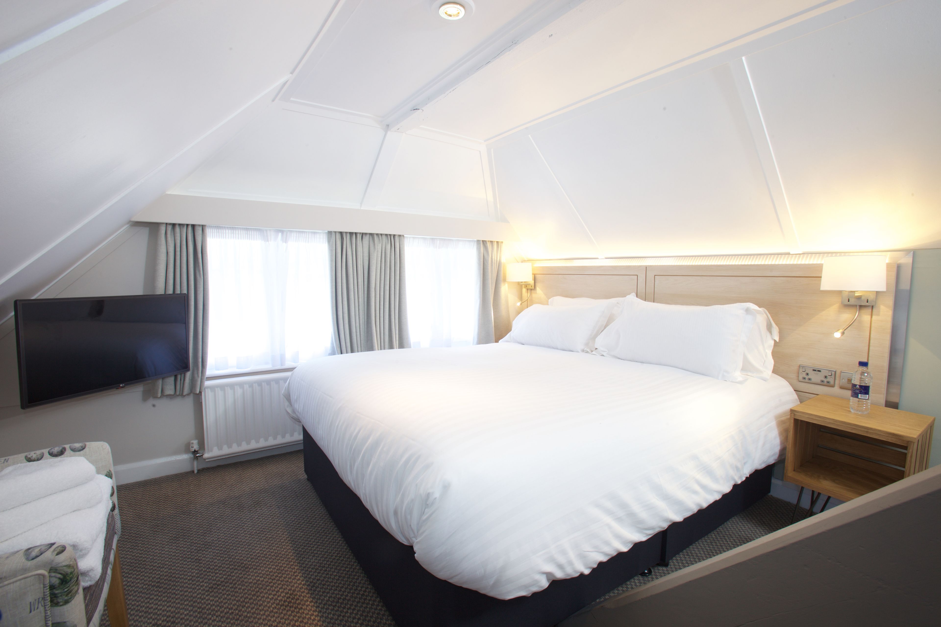 A hotel room with a large bed, a flat-screen TV, a bedside table with a lamp, and a window with curtains.