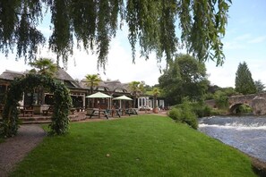 A riverside restaurant with outdoor seating, a thatched-roof building, and a bridge in the background. - Fisherman's Cot, Tiverton by Marston's Inns (Tiverton)