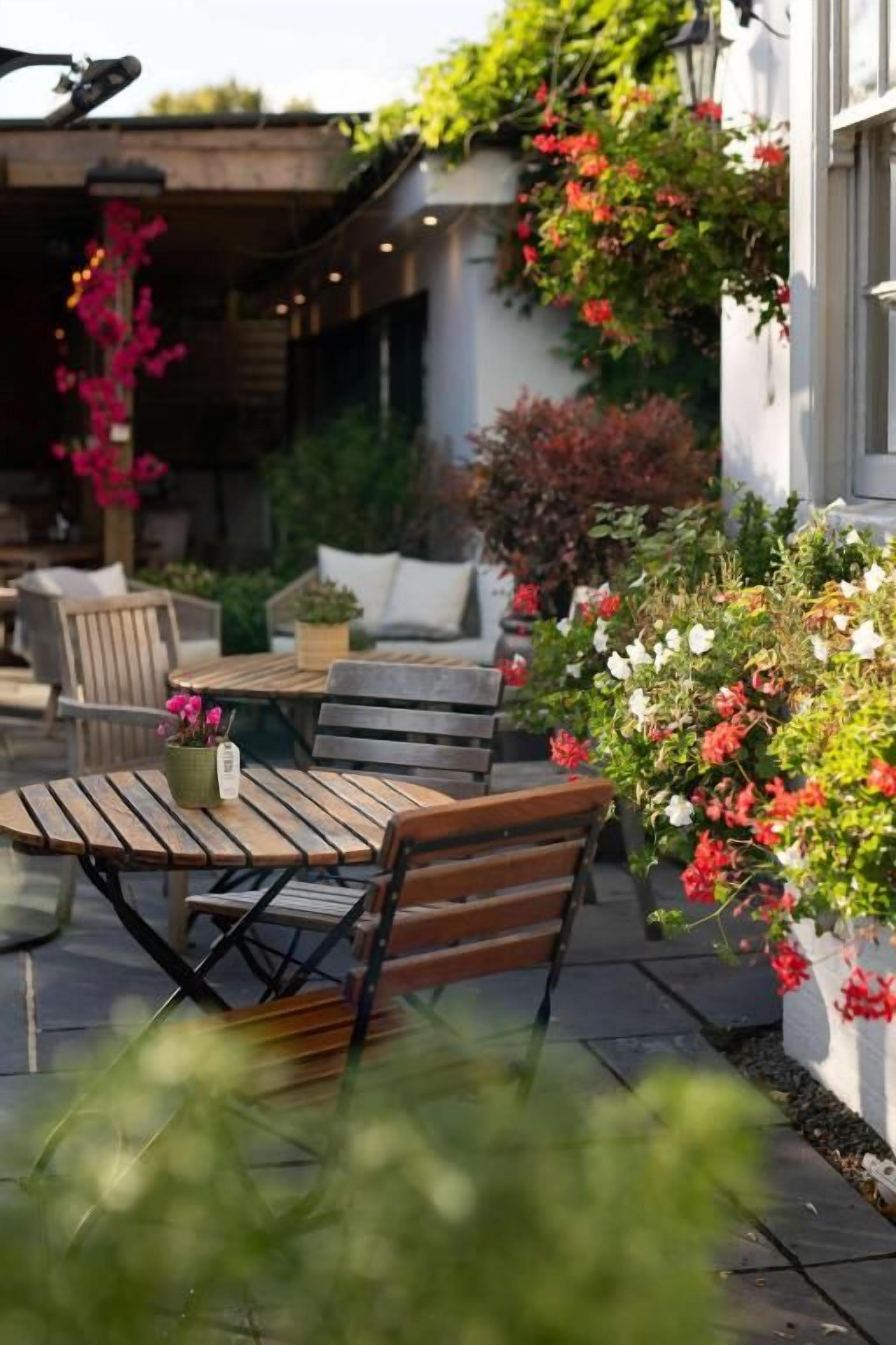 Eine Terrasse im Freien mit einem Holztisch und einer Bank, umgeben von Topfpflanzen und blühenden Sträuchern.