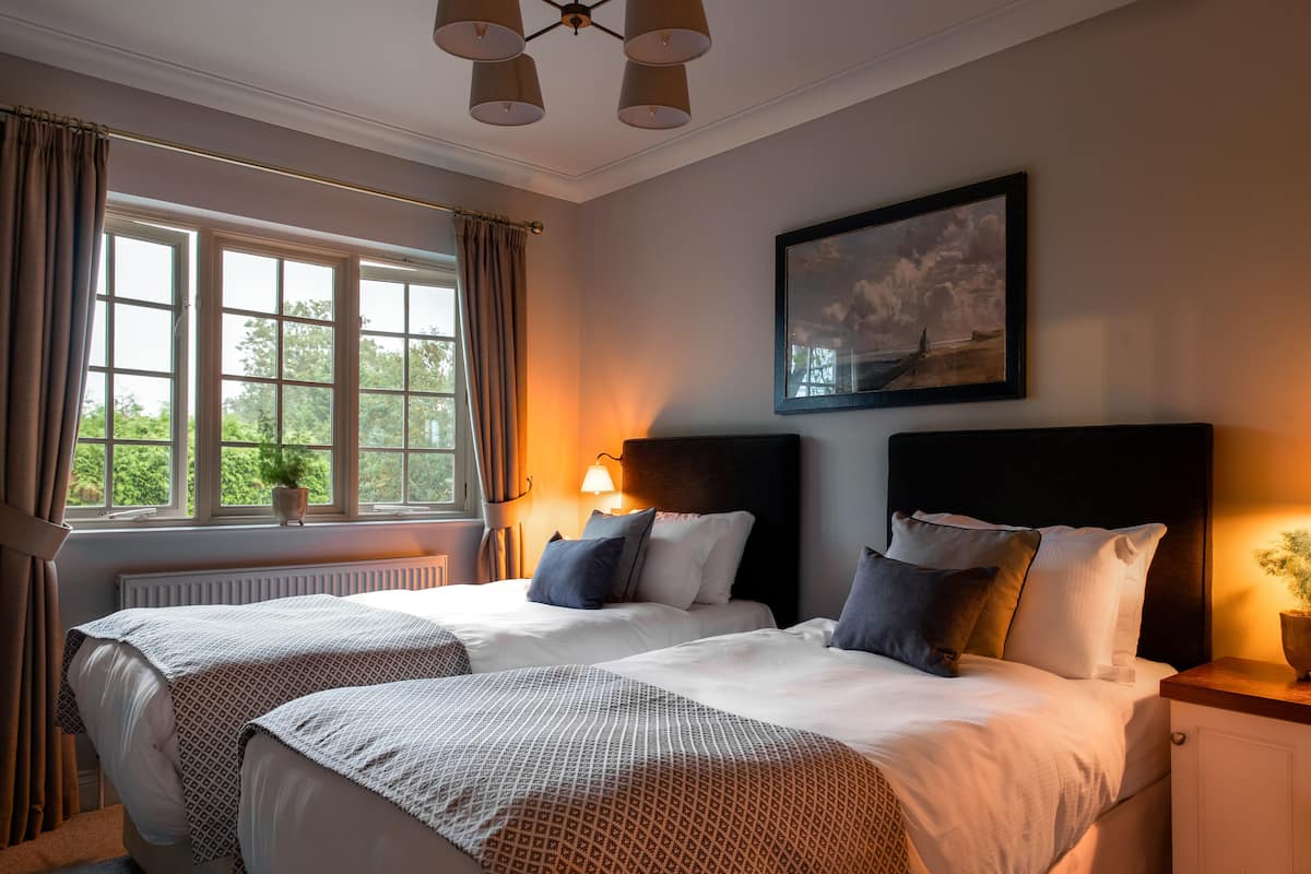 A hotel room with two beds, a large window with curtains, a painting on the wall, and a bedside lamp.