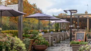 An outdoor seating area with wooden tables and purple umbrellas, surrounded by potted plants and a sign indicating breakfast service. - The Carpenters Arms (Tonbridge)