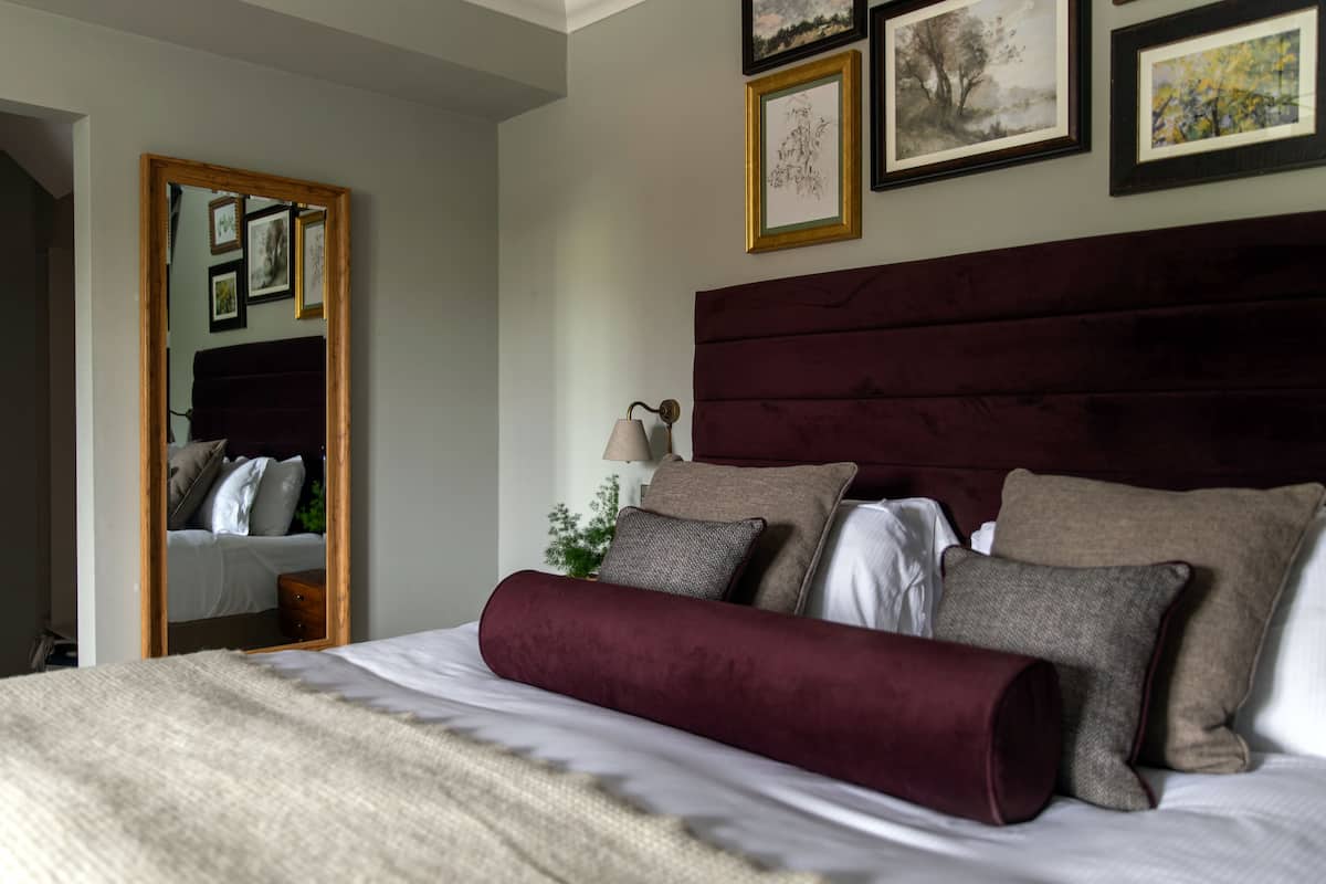 A neatly made bed with a headboard, a bedside lamp, and a mirror reflecting a bedroom with a wardrobe and framed pictures on the wall.