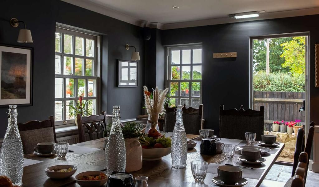 A dining area with a long table set for a meal, large windows, and a view of the outdoors.