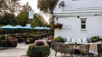 An outdoor seating area with wicker furniture and potted plants, a white building with a sign, and umbrellas.