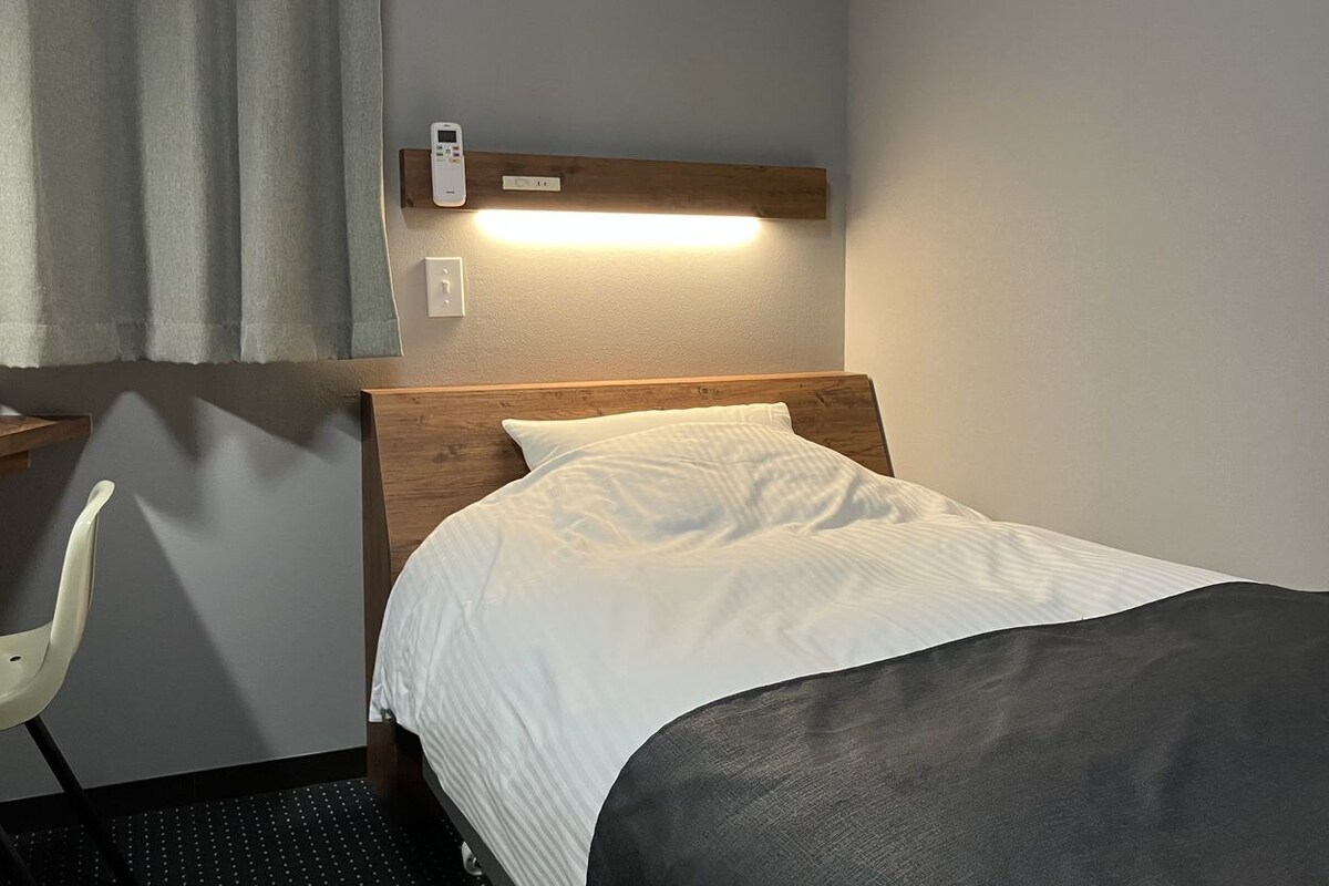 Una habitación de hotel con cama, una silla, una mesita de noche, aire acondicionado instalado en la pared y una lámpara.