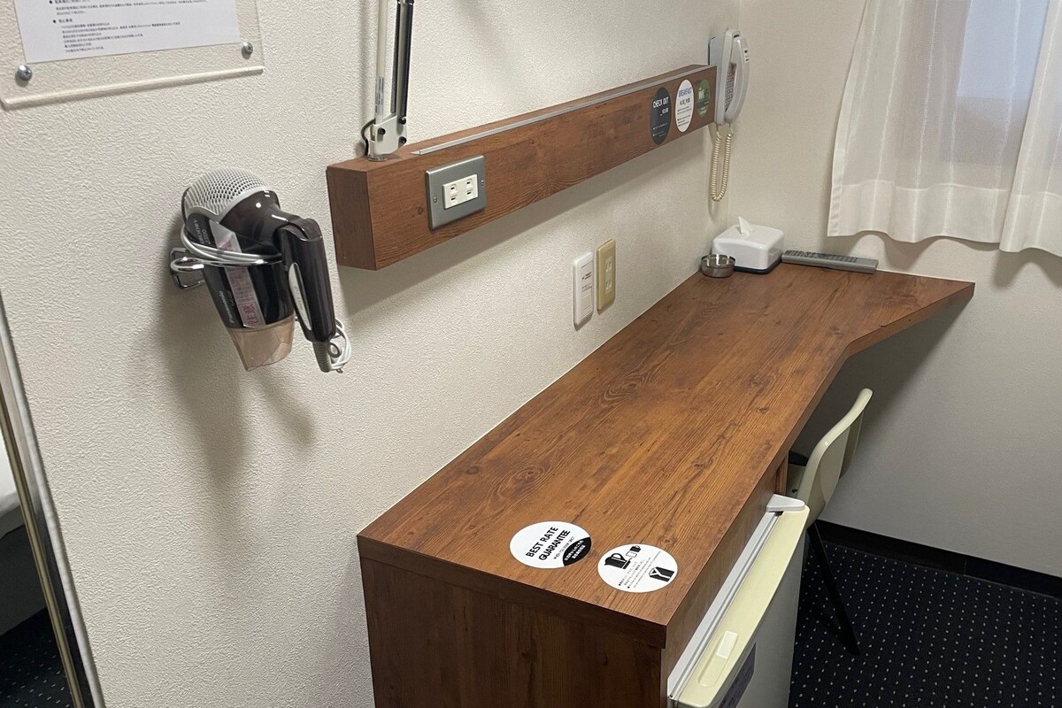 Habitación de hotel pequeña con un escritorio de madera, un teléfono, un cesto de basura y una ventana con cortina translúcida.