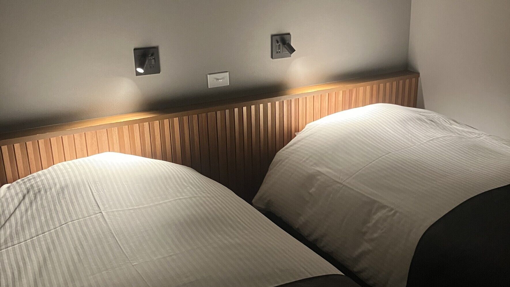 Habitación de hotel con dos camas, cabecero de madera e interruptores de luz fijados a la pared.