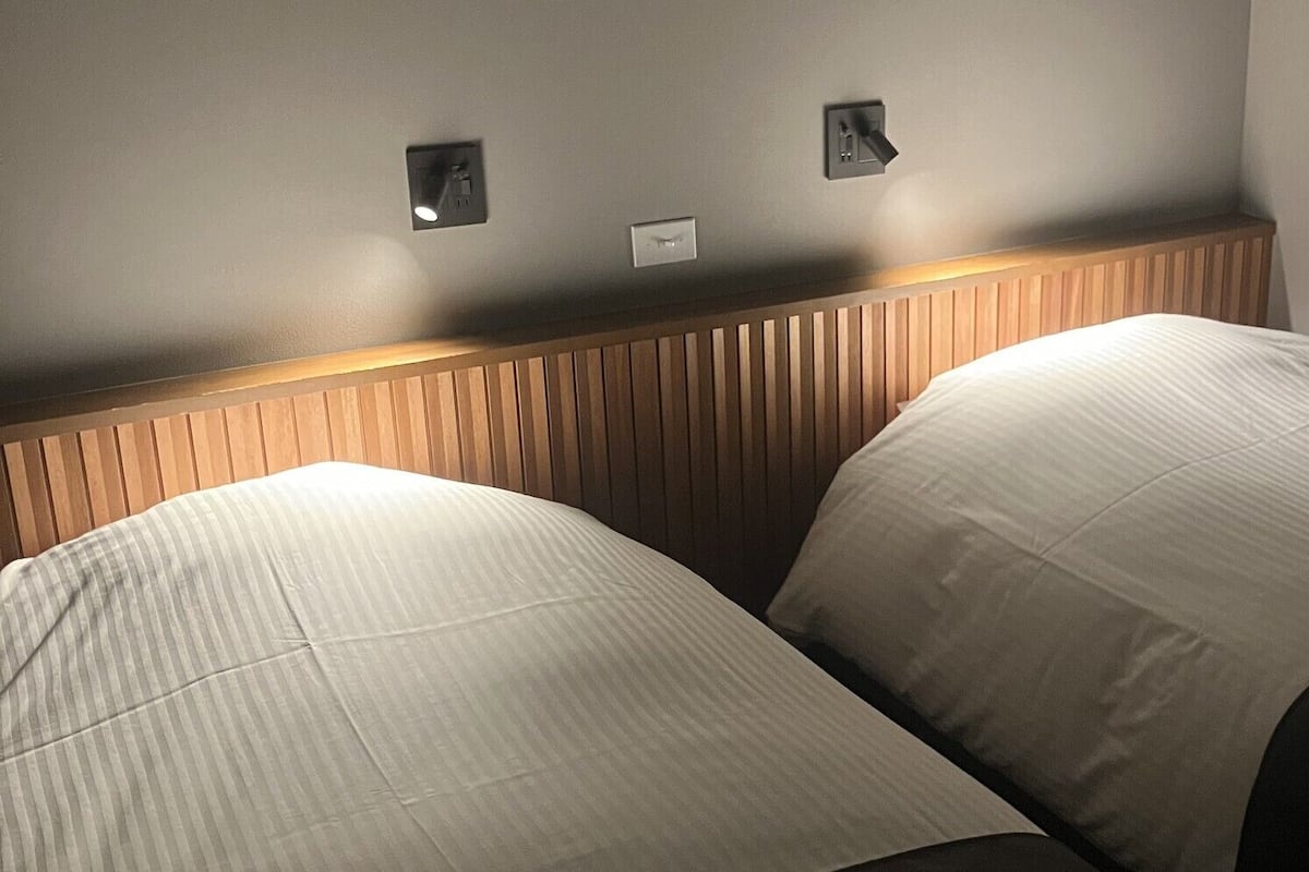 A hotel room with two beds, wooden headboard, and wall-mounted light switches.