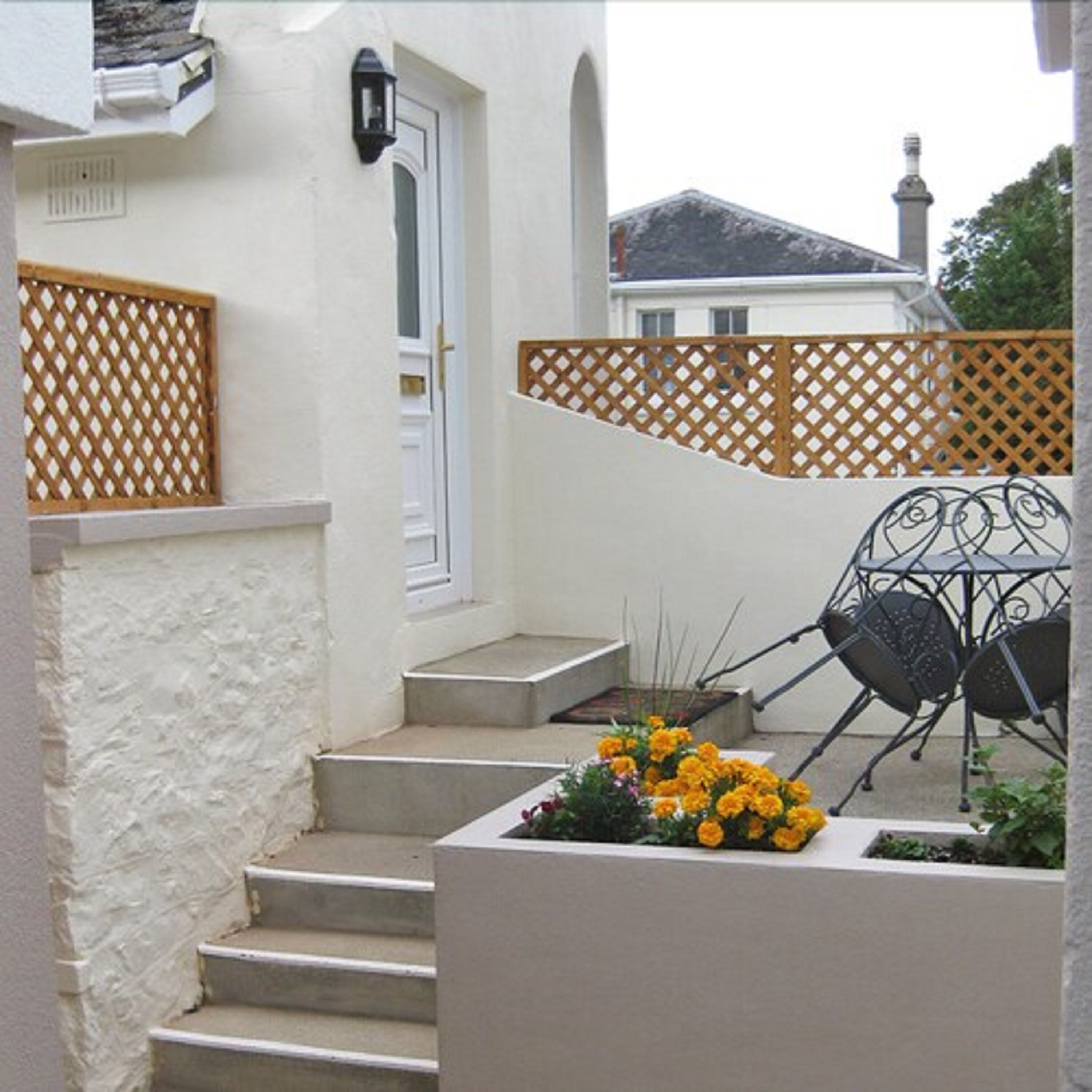 A set of concrete steps leading to a white door, a wooden lattice fence, and a metal outdoor chair with a planter of yellow flowers.