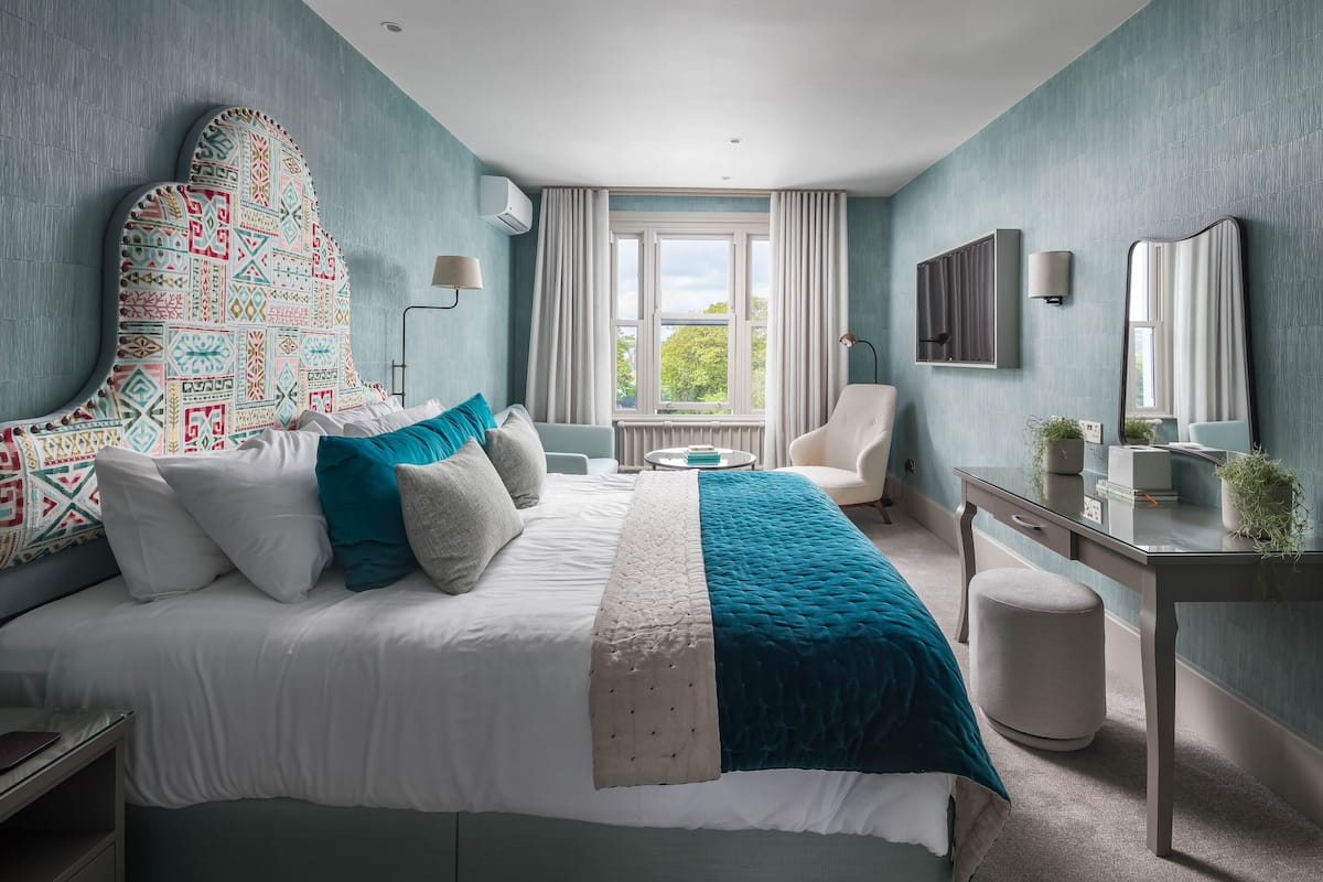 A bedroom with a large bed, a desk, a chair, a window with curtains, and a mirror on the wall.