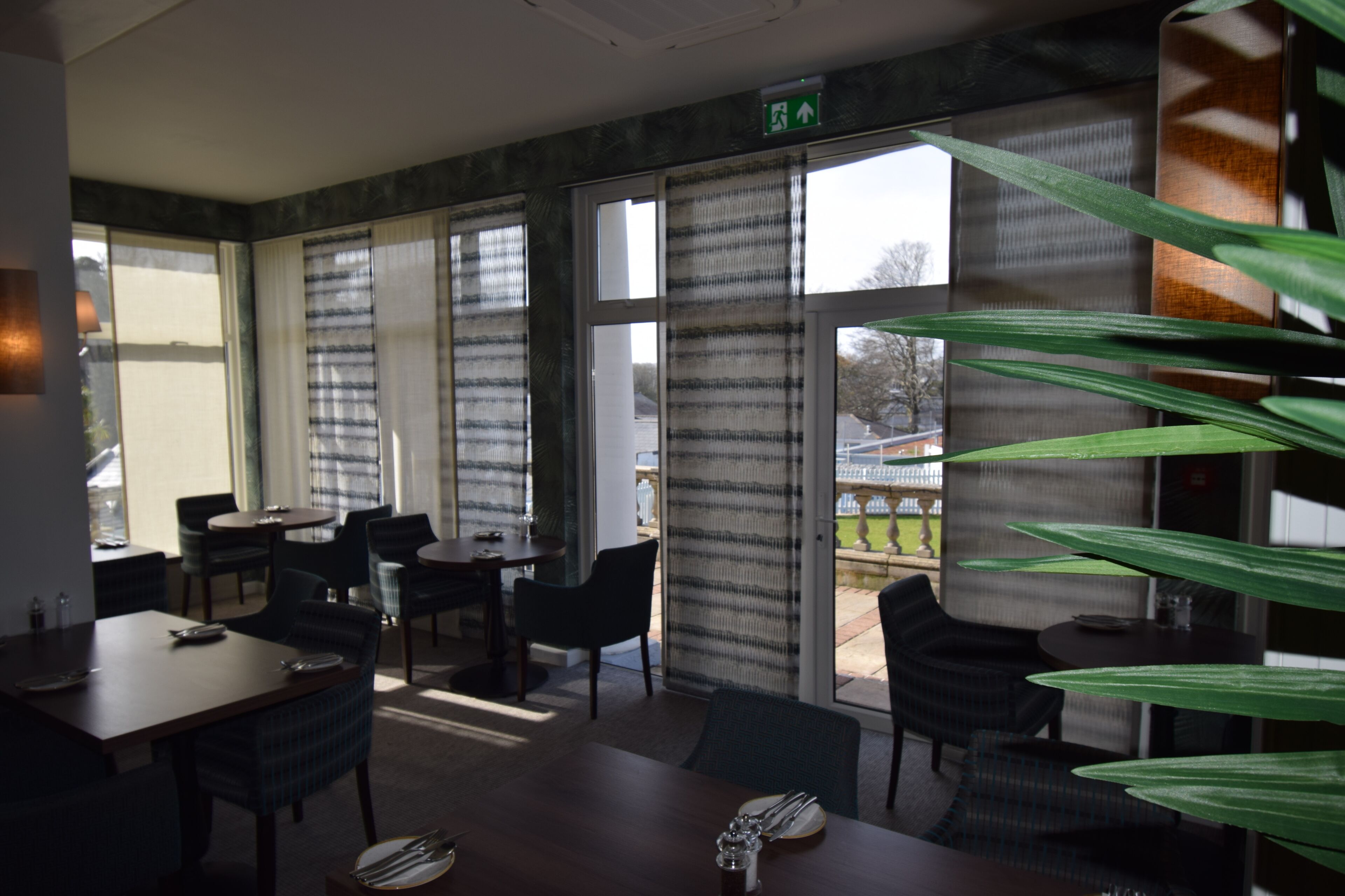 A dining area with tables and chairs, large windows with curtains, and a view of the outdoors.