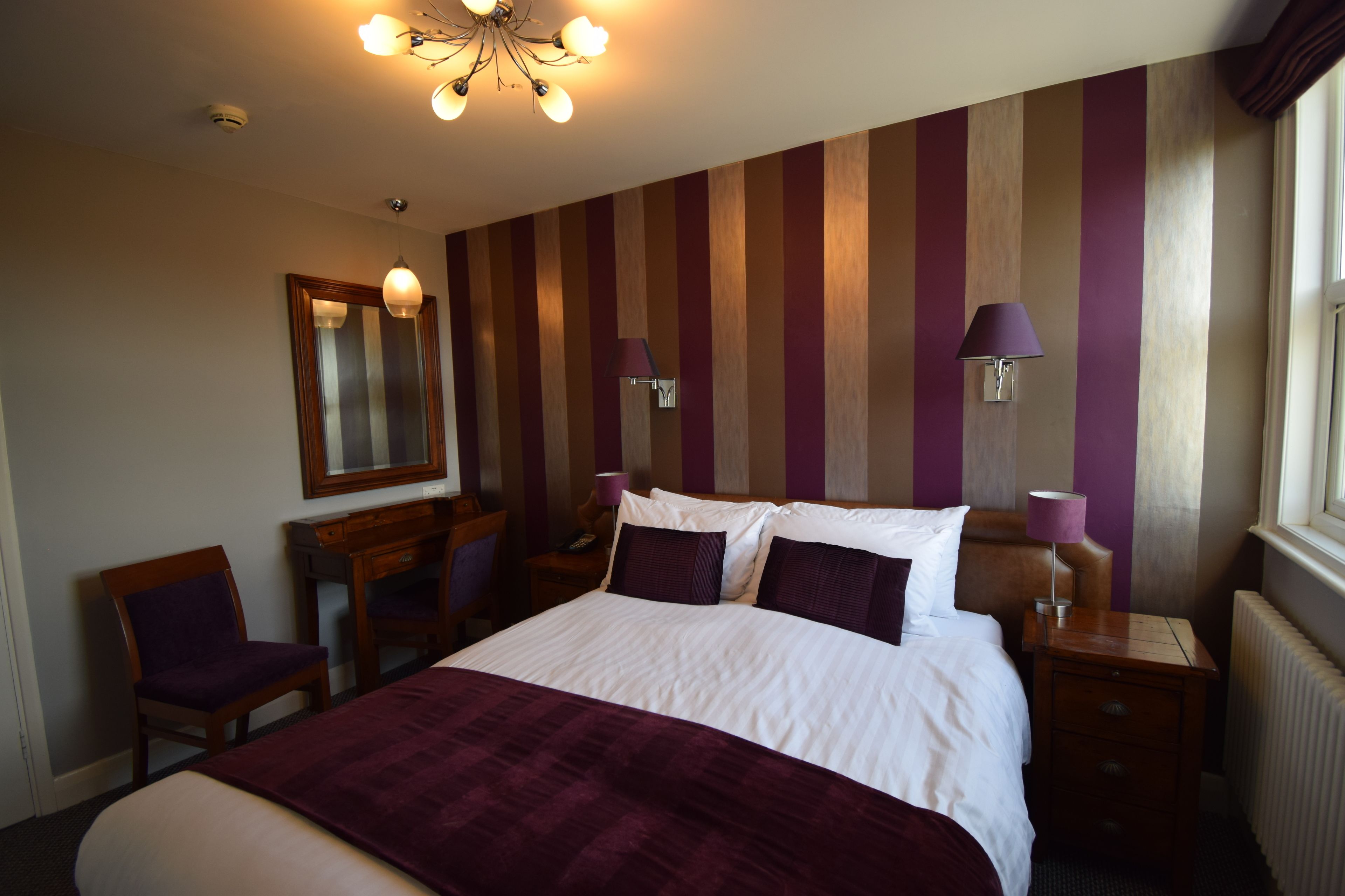 A hotel room with a large bed, a desk, a chair, a nightstand, a lamp, and a window.